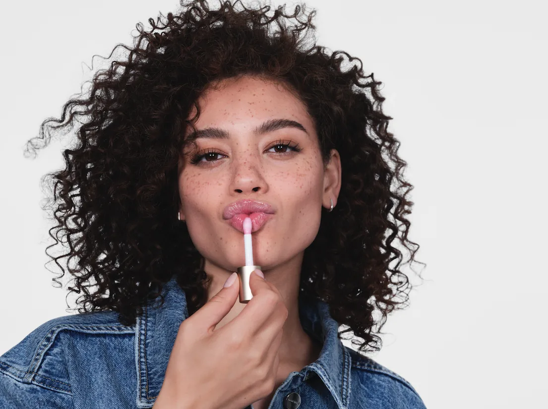 bareMinerals Bare Your Beauty campaign still: curly-haired model in a denim jacket applying lip color