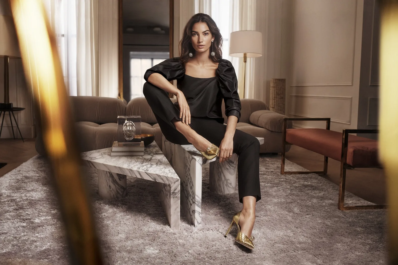 Jimmy Choo FW18 campaign still: Lily Aldridge in black sits on a marble table wearing gold heels