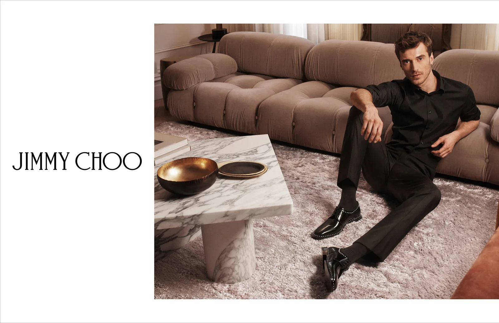 Jimmy Choo FW18 print ad layout: a man in black on a beige sofa wearing patent shoes, with the wordmark