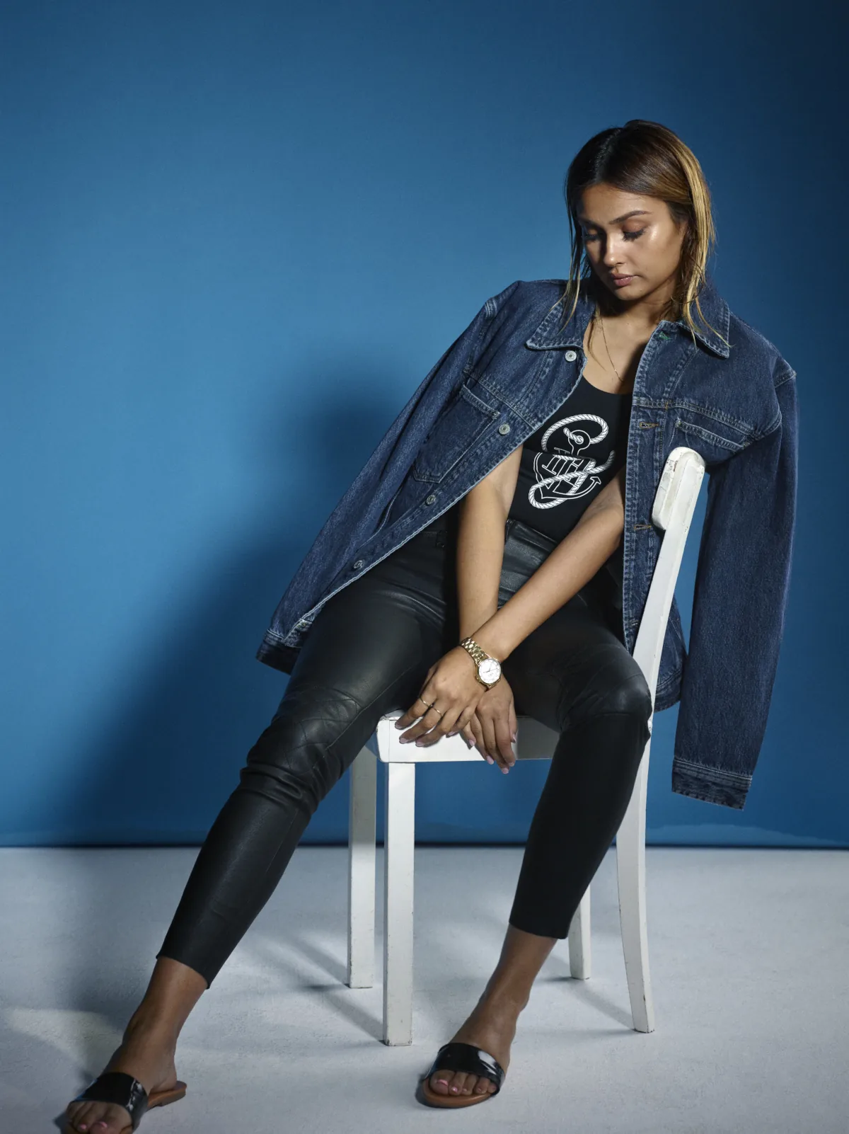Influencer in a denim jacket and leather trousers seated on a white chair against a blue backdrop, Berlin