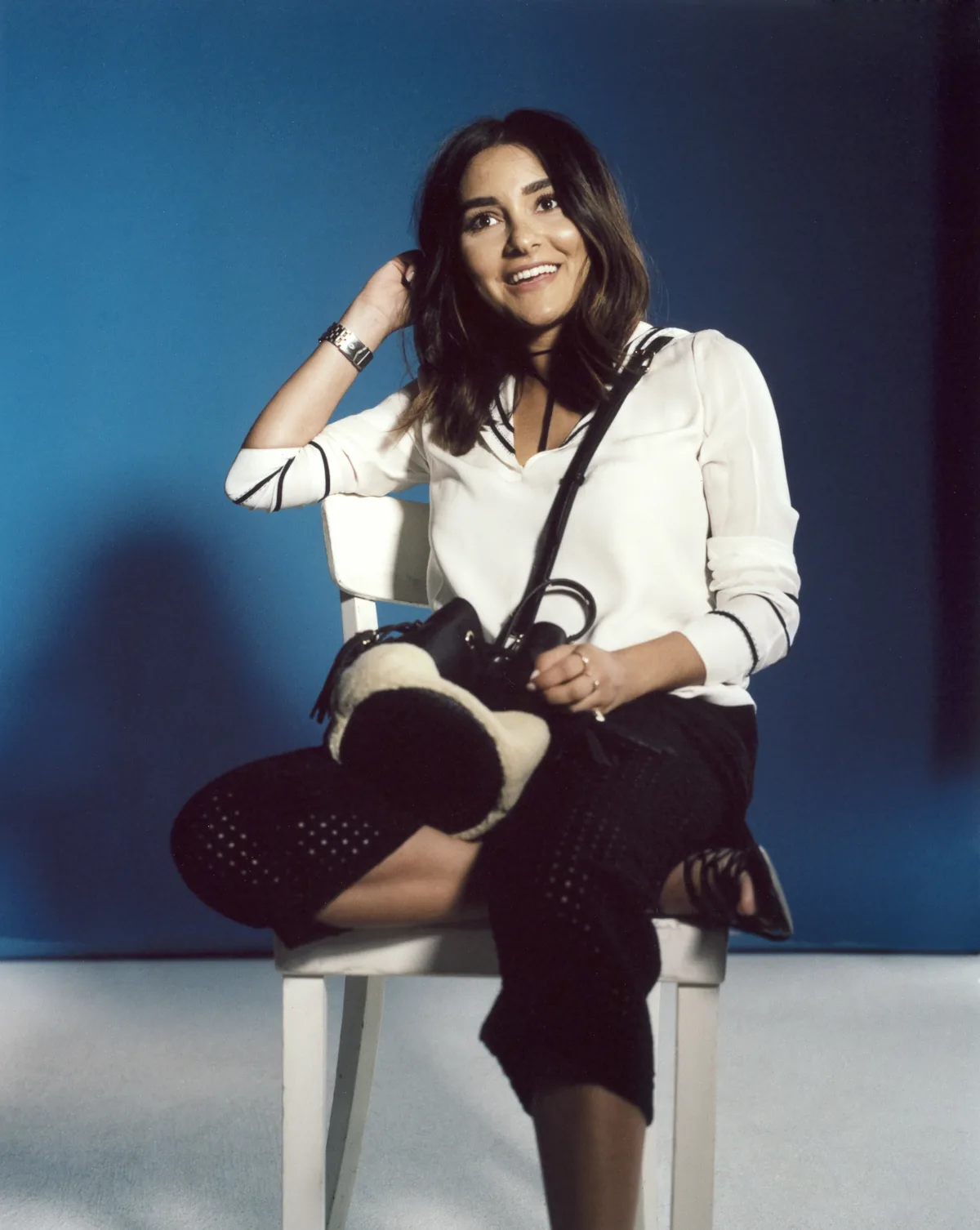 Influencer in a white blouse seated on a white chair with a handbag against a blue backdrop, Berlin
