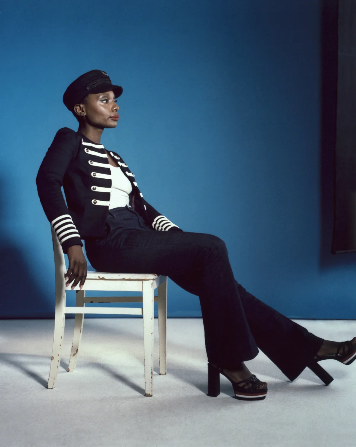 Influencer in a captain's hat and military-striped jacket leaning back on a white chair, blue backdrop
