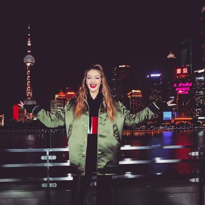 Gigi Hadid in a green bomber jacket with arms open in front of the Shanghai skyline at night