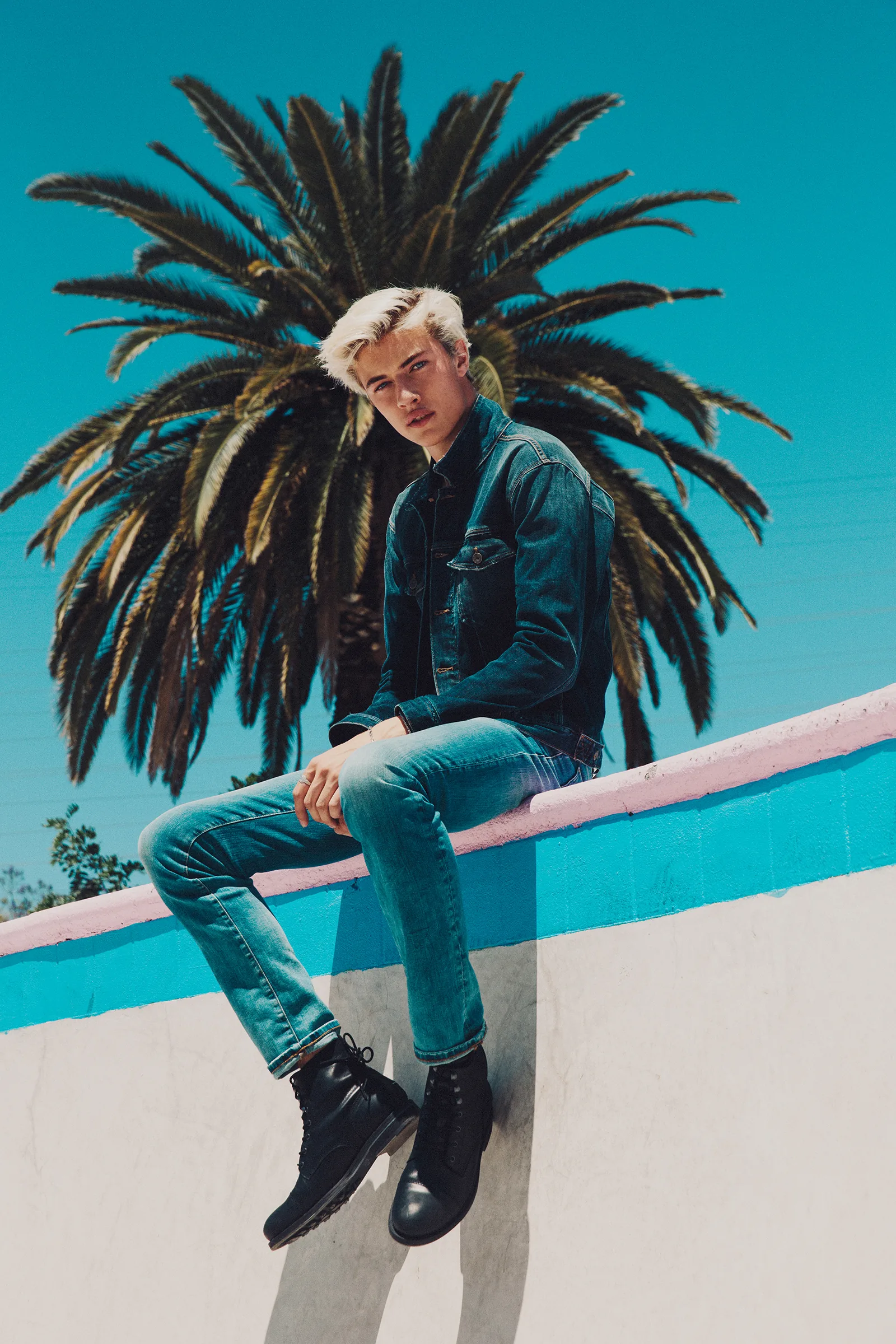 Lucky Blue Smith in denim sitting on a wall under a palm tree and blue sky, shot by Bryant Eslava