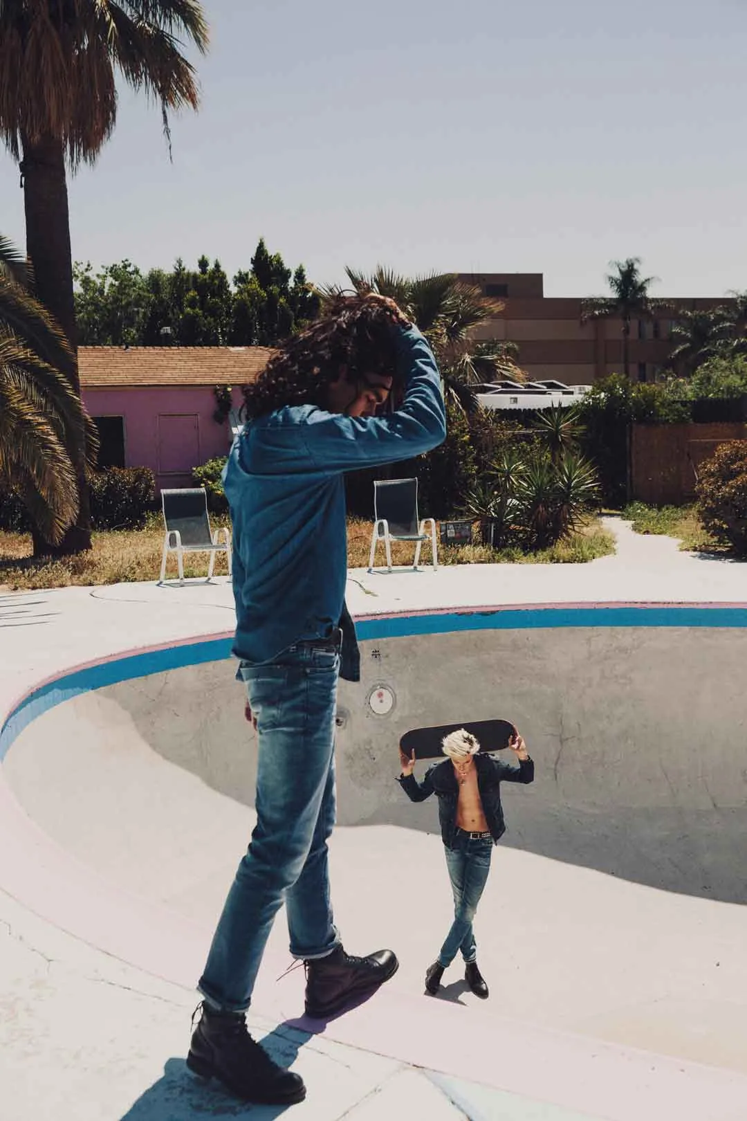 Skater in denim holding a board at the edge of an empty pool with palm trees, shot by Bryant Eslava