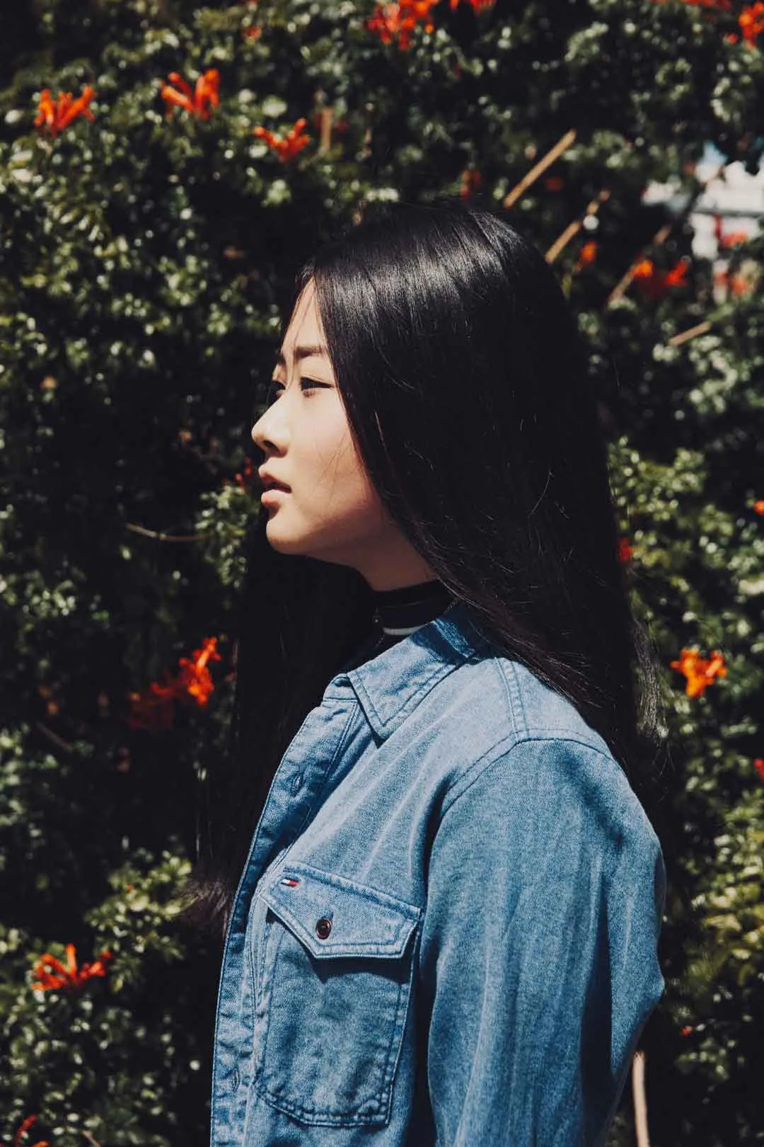 Profile of a woman with long dark hair in a denim shirt beside orange flowers, shot by Bryant Eslava