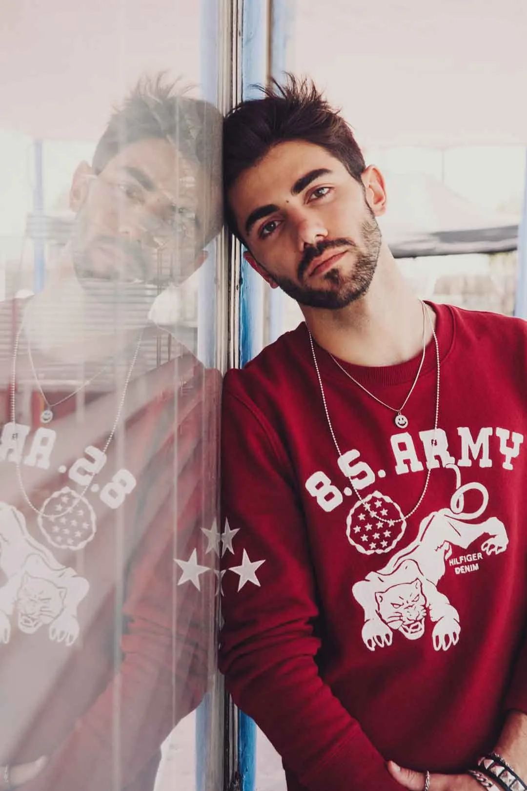 Man in a red B.S. Army sweatshirt leaning against a mirrored window, shot by Bryant Eslava