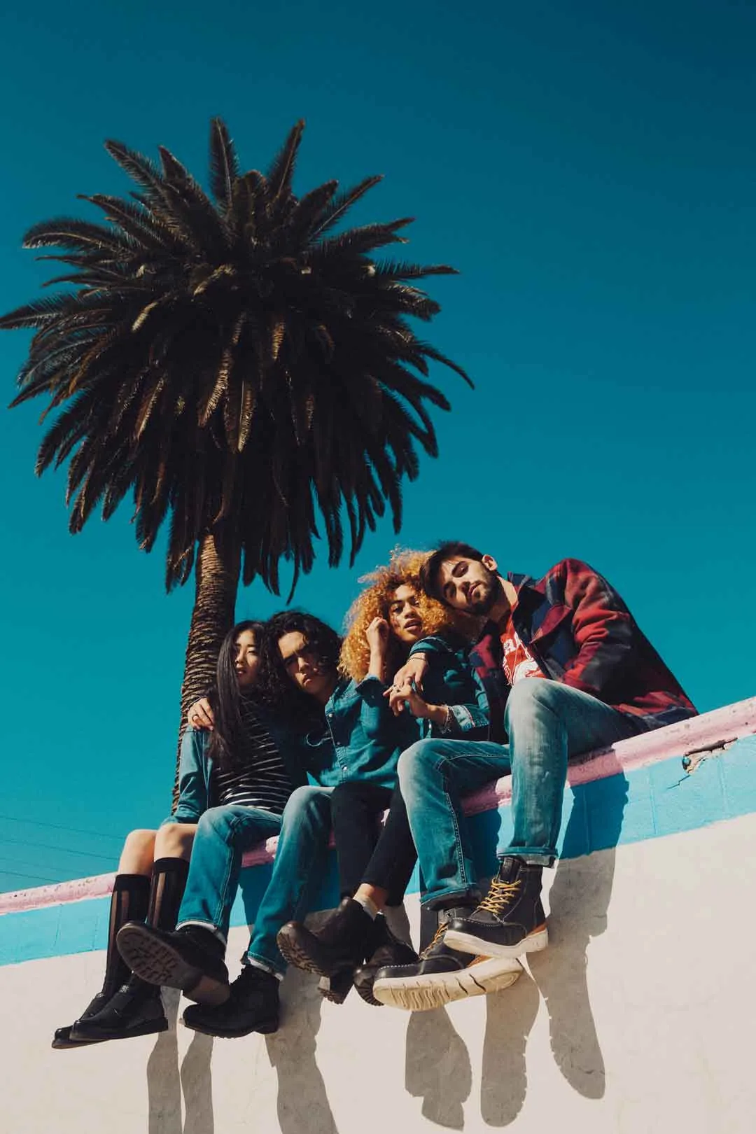 Group of friends in denim seated on the rim of an empty pool beneath a palm tree, shot by Bryant Eslava