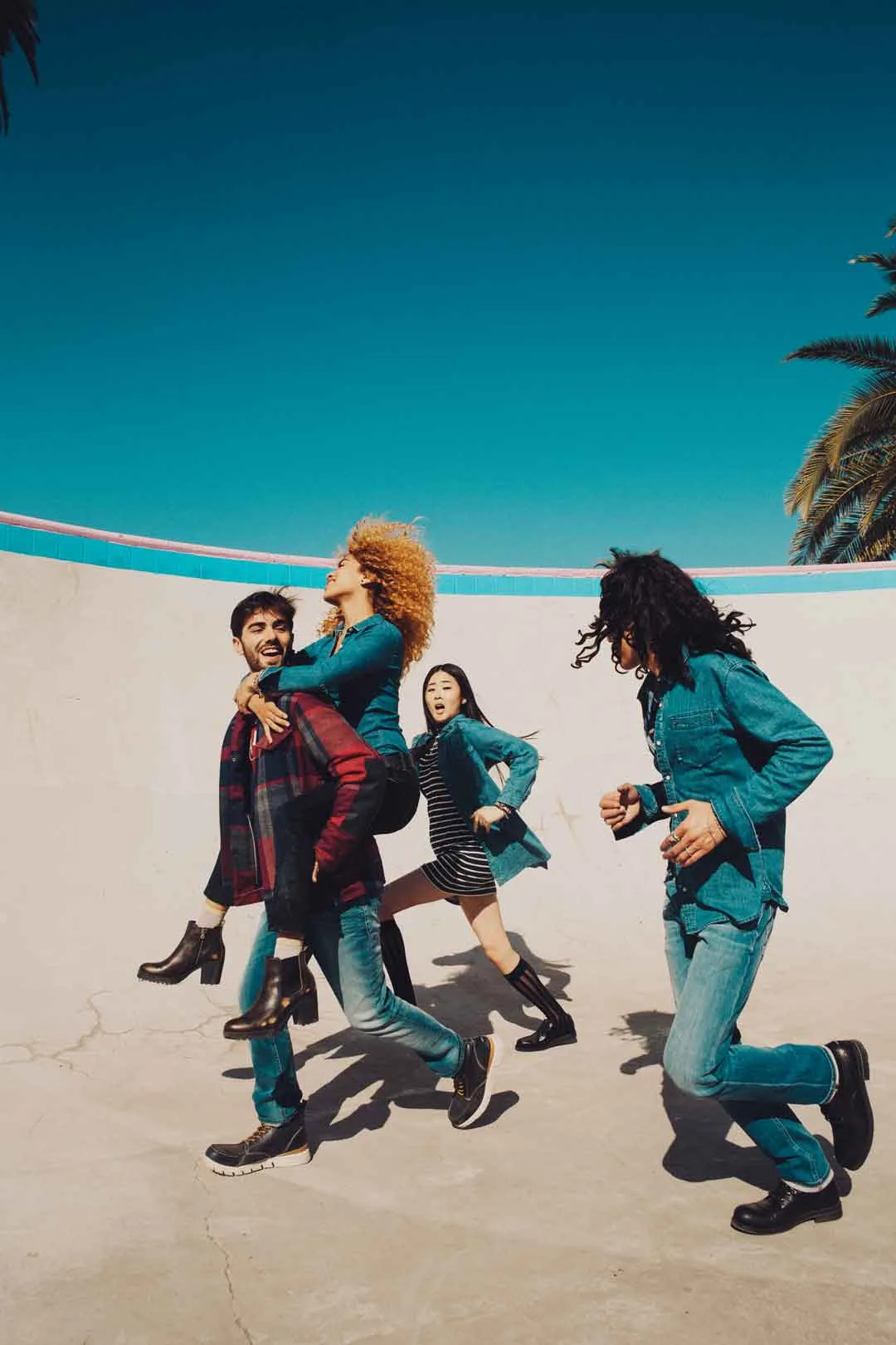 Three friends in denim run across an empty pool, one carried piggyback, shot by Bryant Eslava