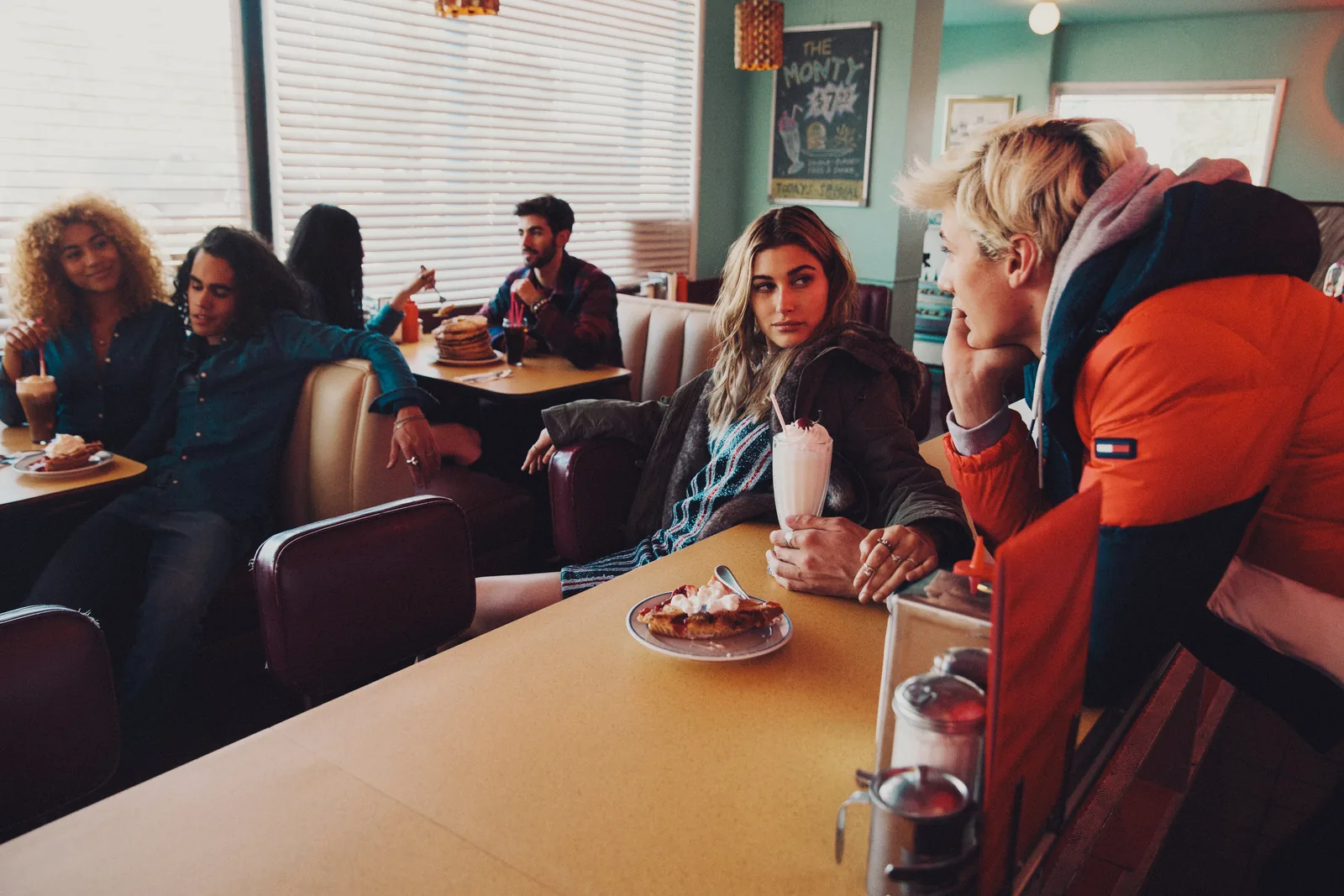Friends in a diner booth while Lucky Blue Smith stands in an orange puffer, shot by Bryant Eslava