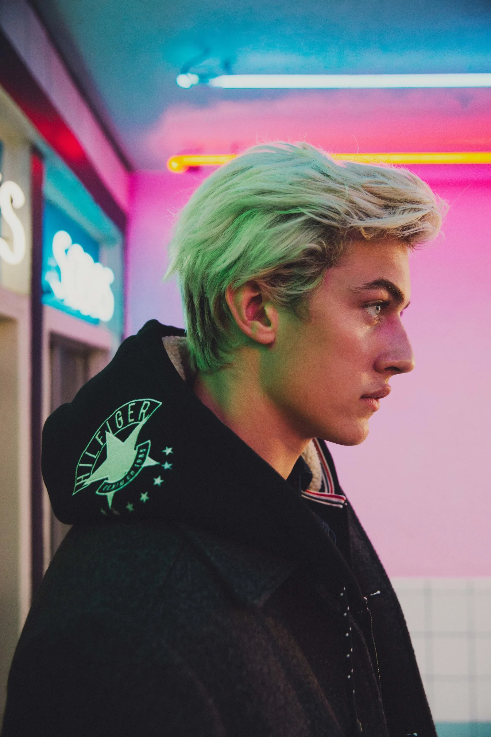 Lucky Blue Smith in profile in a Hilfiger hoodie under a neon Sodas sign, shot by Bryant Eslava