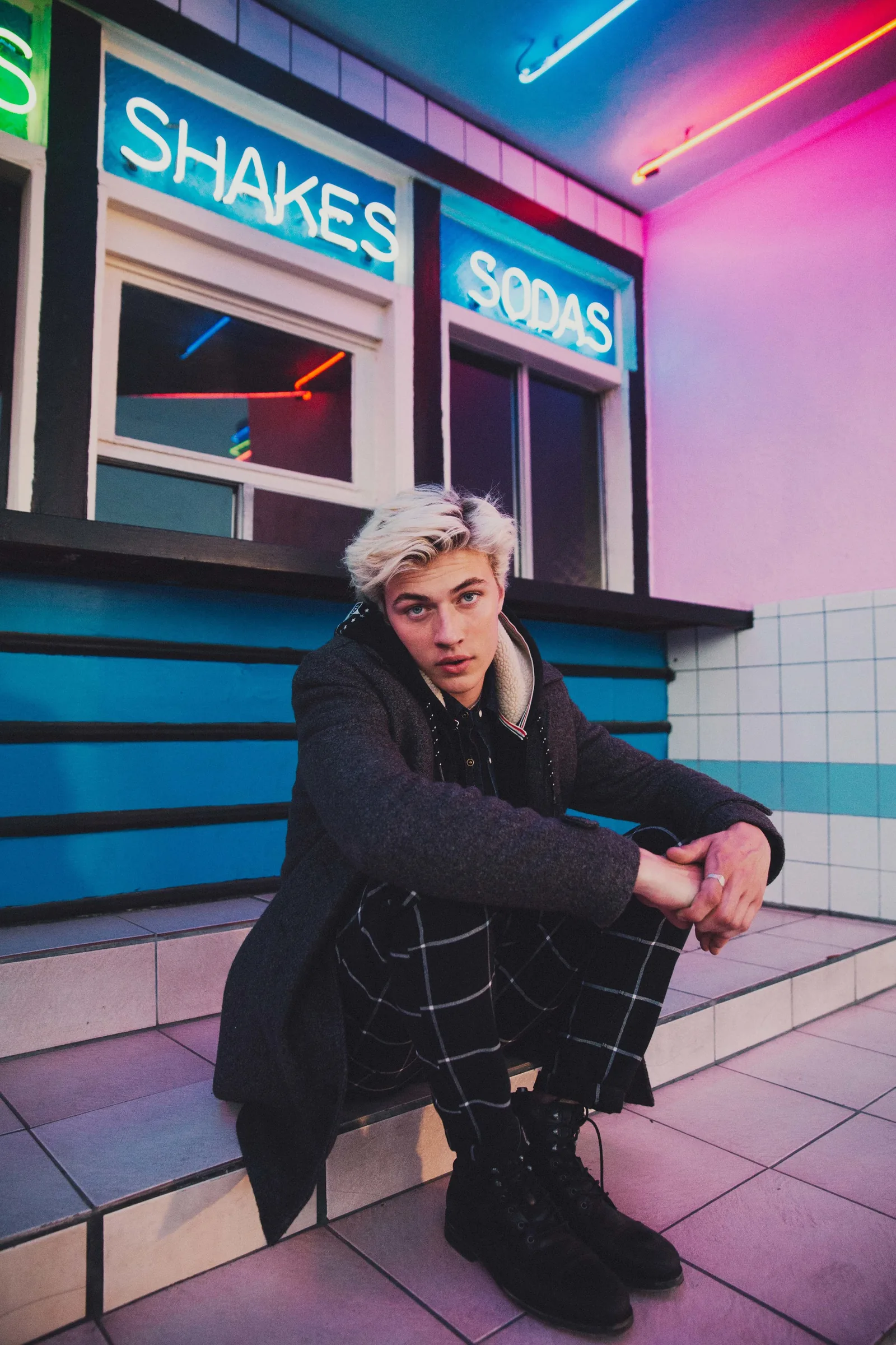 Lucky Blue Smith seated on tiled steps under Shakes and Sodas neon signs, shot by Bryant Eslava