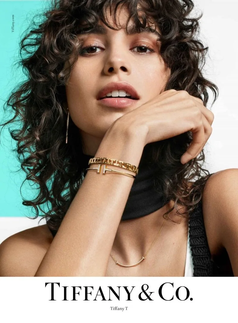 Tiffany T 2019 ad: a curly-haired model rests her chin on her hand wearing a gold T bracelet