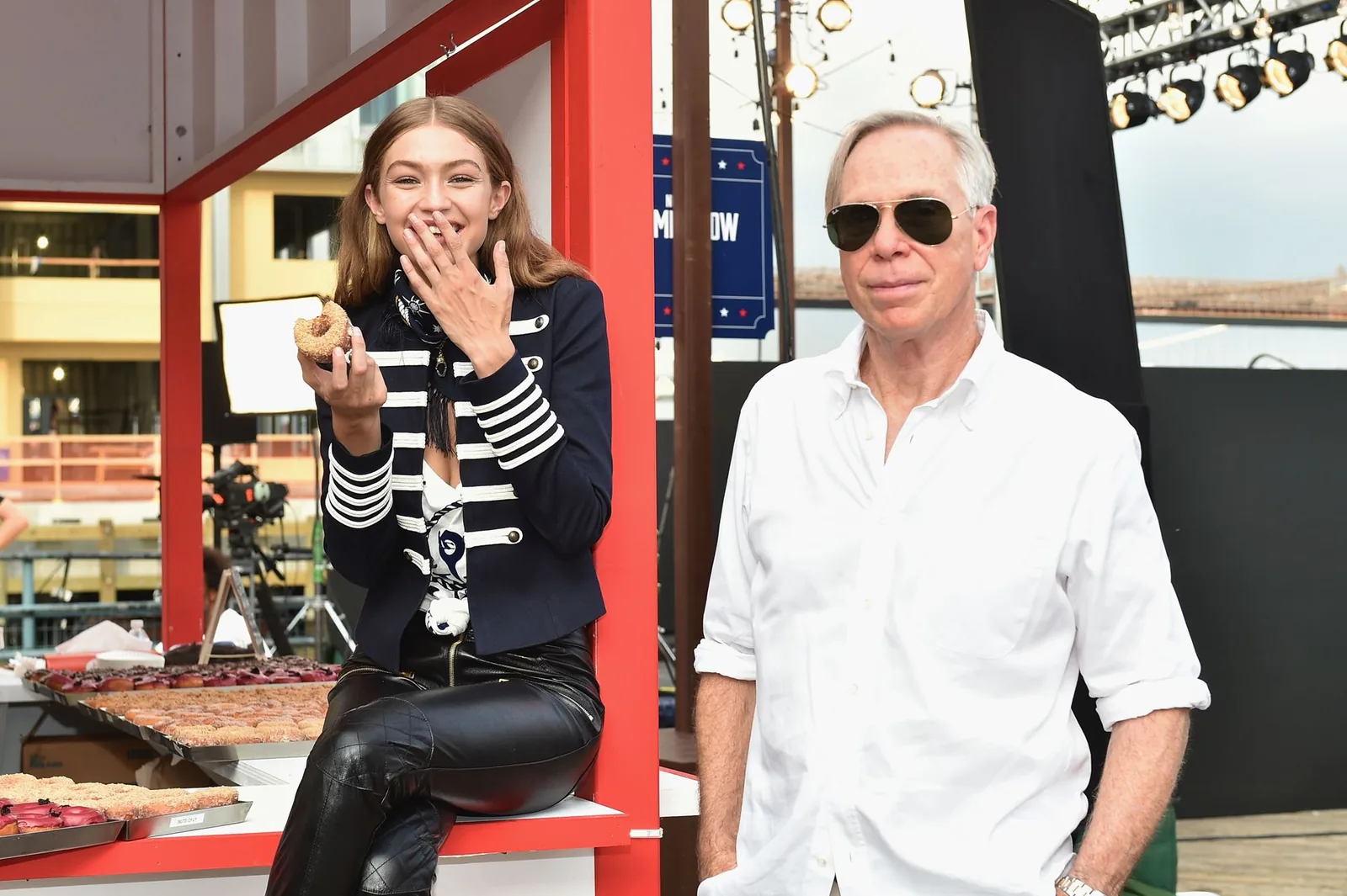 Gigi Hadid eats a donut beside Tommy Hilfiger at the Tommy Pier donut stand